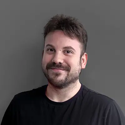 Carlos Moreno, sonriente, con barba y bigote, lleva una camiseta negra y está posando frente a un fondo gris oscuro.